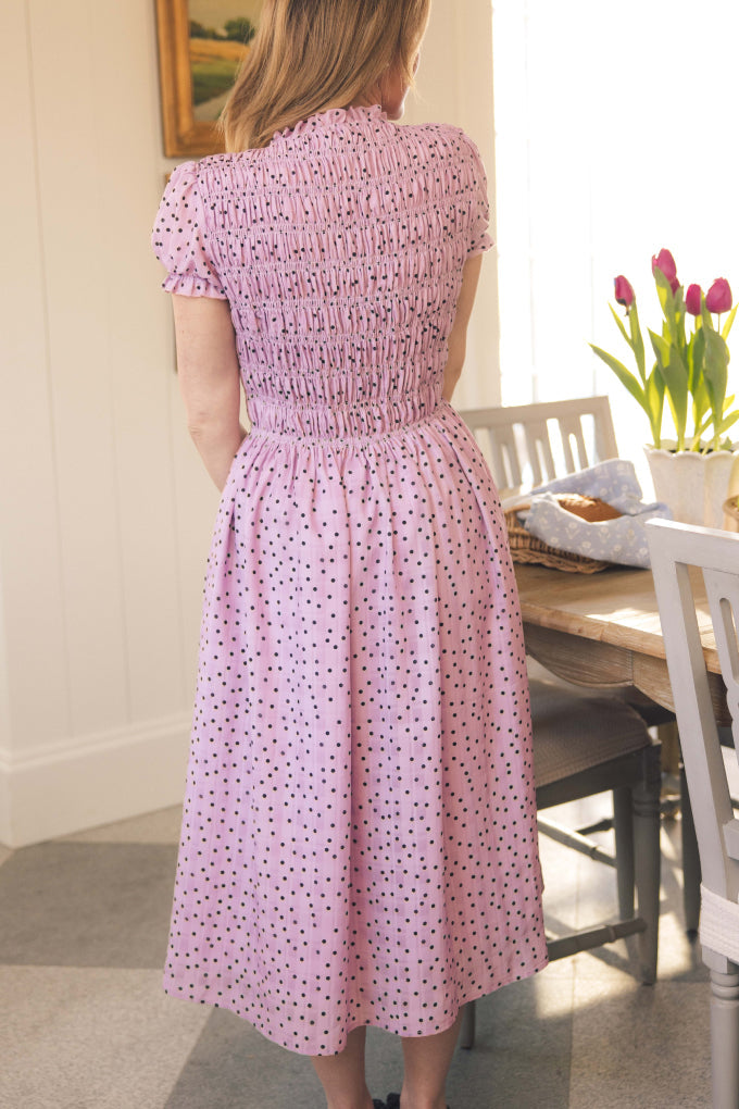 A woman stands indoors with her back to the camera, wearing the Ivy City Co Marcell Dress in Purple Polka Dot. A wooden table with pink tulips is nearby; this maternity-friendly dress features playful puff sleeves and a chic midi length.