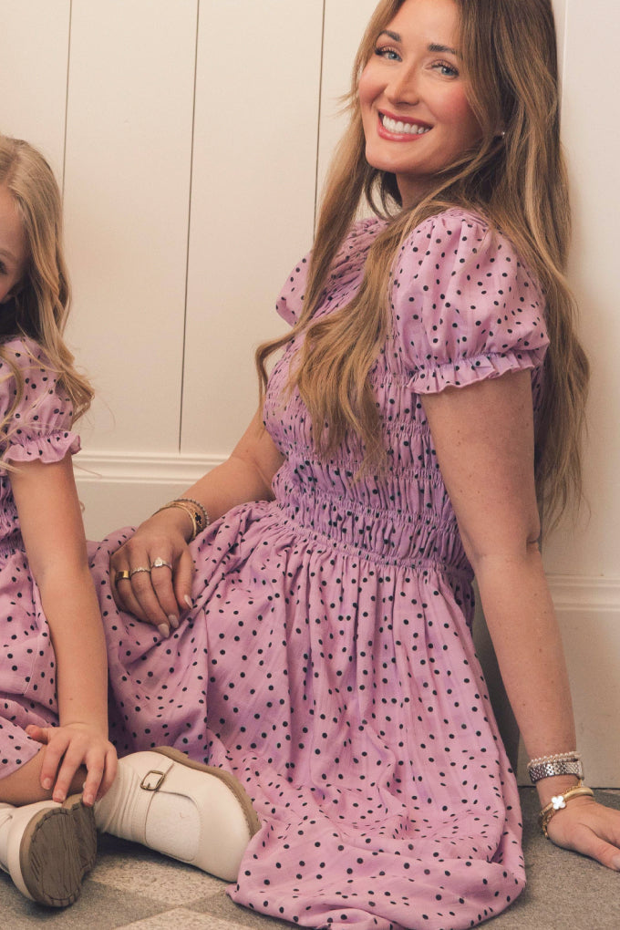 A woman with long brown hair wears the Ivy City Co Marcell Dress in Purple Polka Dot, a maternity-friendly puff sleeve midi dress, as she sits on the floor smiling. A young child in a matching dress is partially visible beside her.