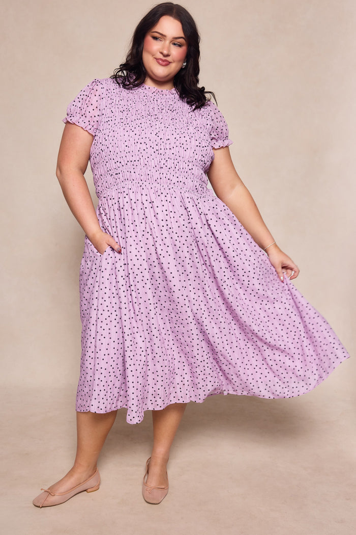 A woman poses in the Ivy City Co Marcell Dress in Purple Polka Dot, holding out the skirt. She has long dark hair and wears light flats. The plain beige background highlights this elegant, maternity-friendly midi dress.