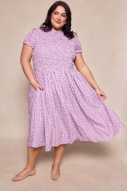 A woman poses in the Ivy City Co Marcell Dress in Purple Polka Dot, holding out the skirt. She has long dark hair and wears light flats. The plain beige background highlights this elegant, maternity-friendly midi dress.