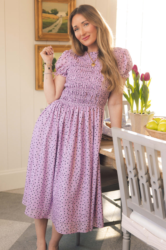 A woman with long hair wearing the Marcell Dress in Purple Polka Dot by Ivy City Co stands beside a dining table adorned with a vase of tulips and a bowl of lemons, sunlight streaming through the window. Paintings and chairs can be seen in the background.