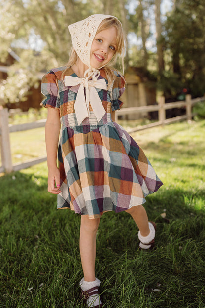 A smiling young girl stands on green grass with a wooden fence and trees in the background, wearing the Ivy City Co Mini Alice Dress in Maize Gingham featuring puff sleeves and a smocked bodice, paired with a lace bonnet.