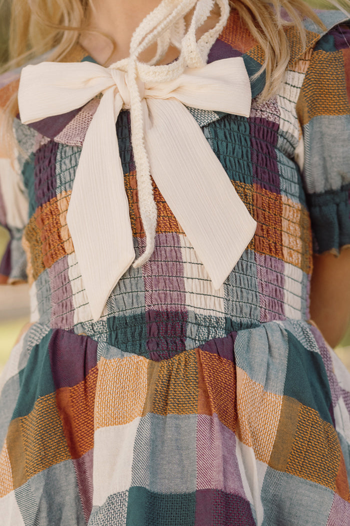 A close-up of a person wearing the Mini Alice Dress in Maize Gingham by Ivy City Co—a vibrant plaid dress with a smocked bodice, charming puff sleeves, and a large cream bow at the neckline.