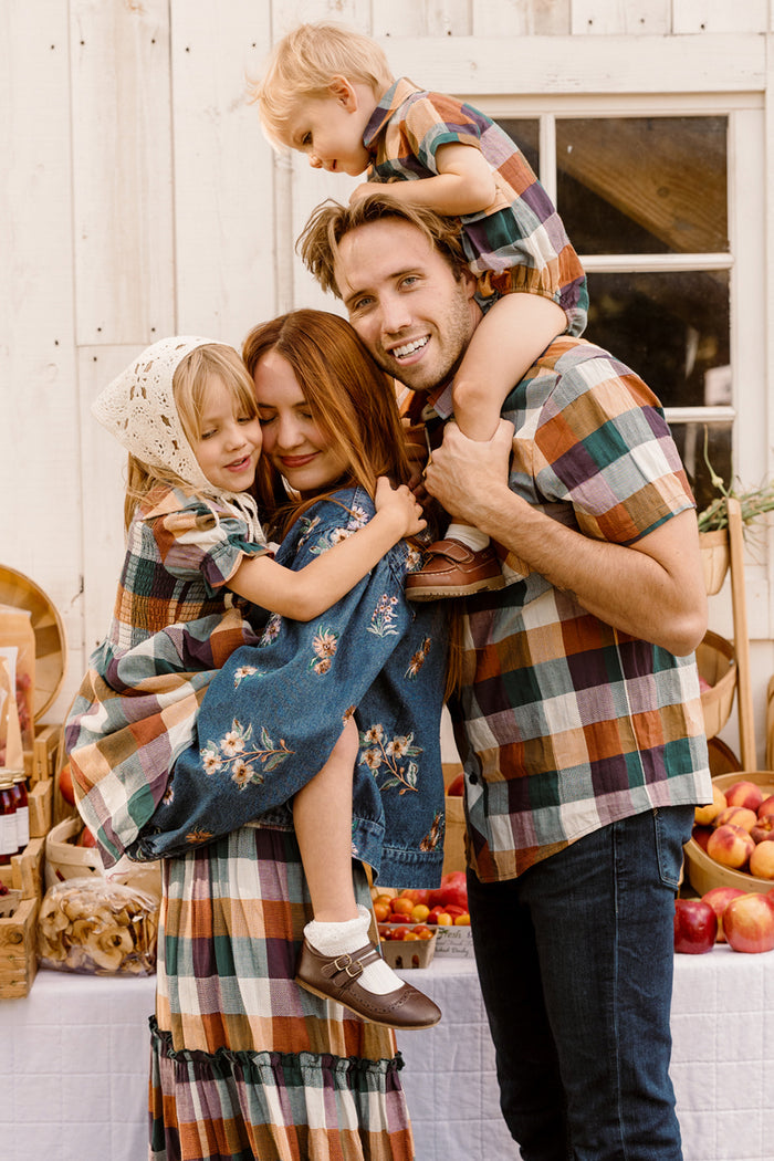 A family of four stands together smiling. The father carries a young child on his shoulders, while the mother holds another child dressed in the Ivy City Co Mini Alice Dress in Maize Gingham, featuring puff sleeves and a smocked bodice. The family wears matching plaid outfits as they pose by a table with fruit and jars, beside a white building.