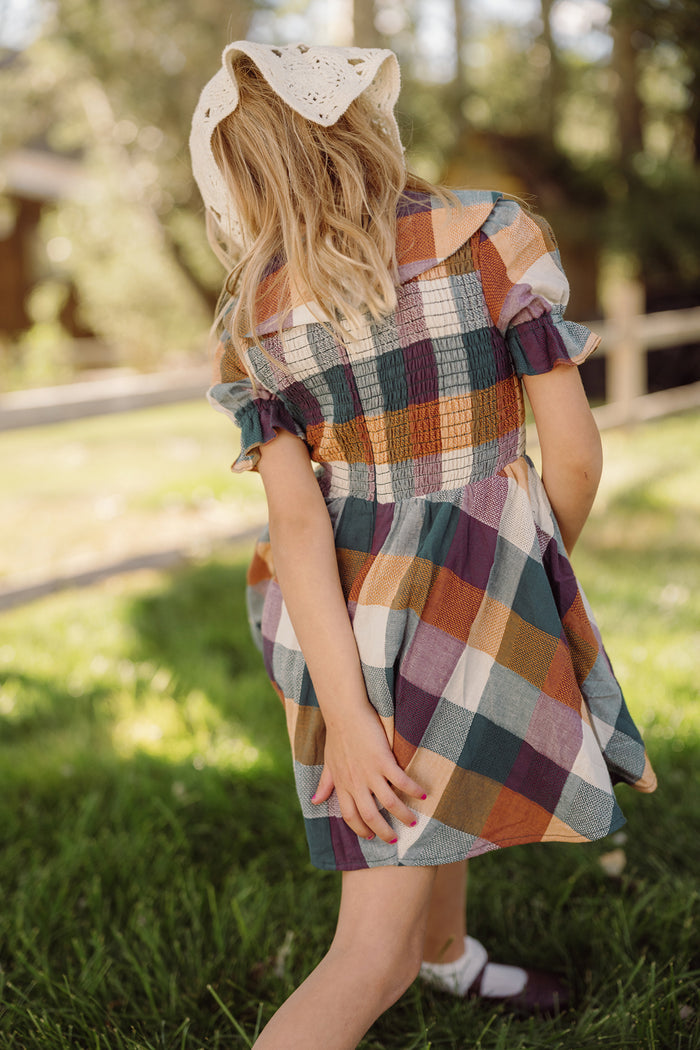 A young girl in the Mini Alice Dress in Maize Gingham by Ivy City Co, featuring puff sleeves and a smocked bodice, stands on green grass facing away, one hand behind her back as sunlight filters through the trees in the background.