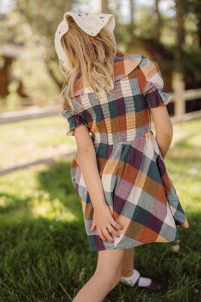 A young girl in the Mini Alice Dress in Maize Gingham by Ivy City Co, featuring puff sleeves and a smocked bodice, stands on green grass facing away, one hand behind her back as sunlight filters through the trees in the background.
