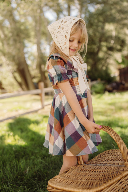A young girl wearing the Ivy City Co Mini Alice Dress in Maize Gingham, featuring a smocked bodice and puff sleeves, stands on grass outdoors holding a wicker basket, with trees and a wooden fence in the background.