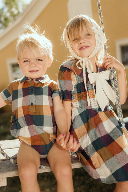 Two young children, both wearing matching plaid outfits, sit together on a wooden swing outdoors. The older child wears the charming Mini Alice Dress in Maize Gingham by Ivy City Co, featuring puff sleeves and a smocked bodice; both children are smiling, holding hands, and enjoying the sunlight.