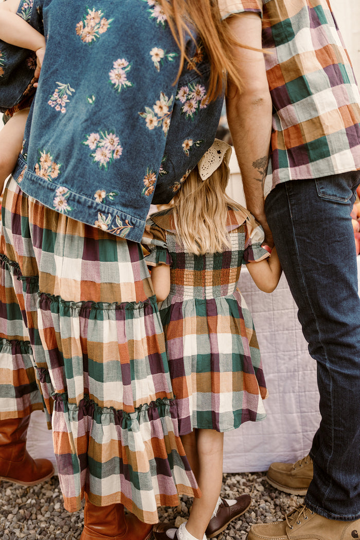 A young girl in an Ivy City Co Mini Alice Dress in Maize Gingham, featuring puff sleeves and a smocked bodice, holds hands with adults in coordinating fall outfits, viewed from behind.
