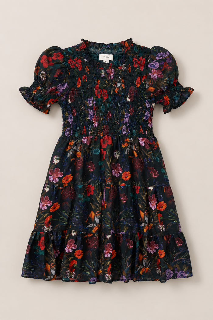 The Mini Cordelia Dress in Evergreen Vivid Meadow by Ivy City Co is a short-sleeved, knee-length dress featuring a smocked bodice, elegant puff sleeves, and a ruffled hem. It showcases a vibrant floral print in red, purple, and green on a black background.