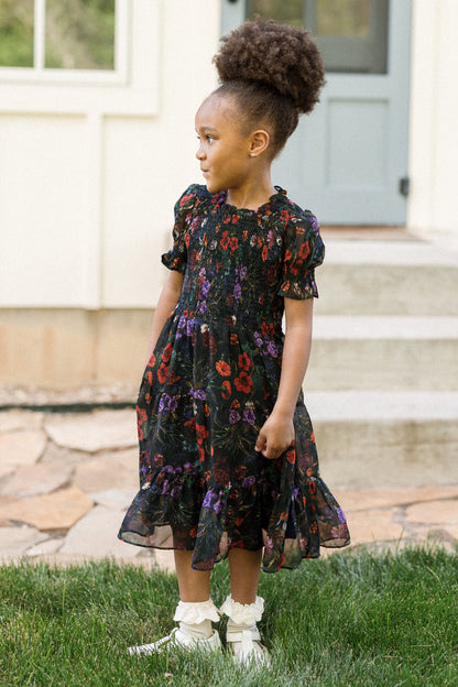 A young girl with a high curly ponytail wears the Ivy City Co Mini Cordelia Dress in Evergreen Vivid Meadow, featuring puff sleeves, along with white shoes and socks. She stands on grass, looking to the side, with stone steps and a house in the background.