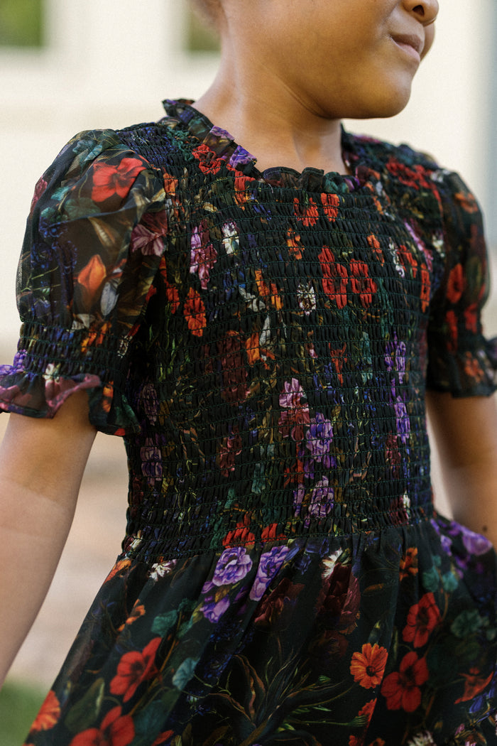 A child wears the Ivy City Co Mini Cordelia Dress in Evergreen Vivid Meadow, which features a black base adorned with vibrant red, purple, and green floral patterns. The dress highlights a smocked bodice and puff sleeves accented with ruffled details. The child is smiling softly, set against a gently blurred background.