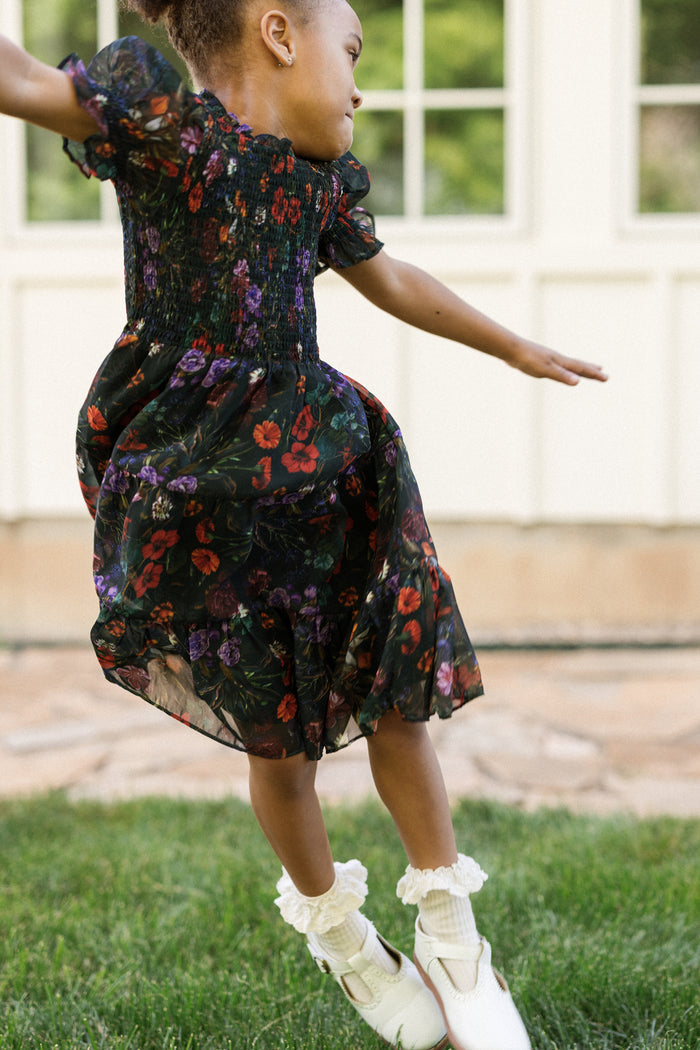 A young girl in the Ivy City Co Mini Cordelia Dress in Evergreen Vivid Meadow, featuring a smocked bodice and puff sleeves, jumps playfully on grass in front of a light-colored building, her white shoes perfectly completing the look.