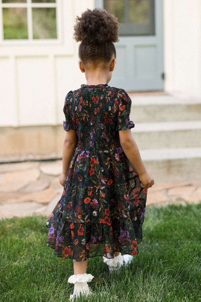 A young girl with curly hair, wearing the Ivy City Co Mini Cordelia Dress in Evergreen Vivid Meadow with puff sleeves and white socks, stands on grass facing away from the camera in front of a house with stone steps and a light blue door.