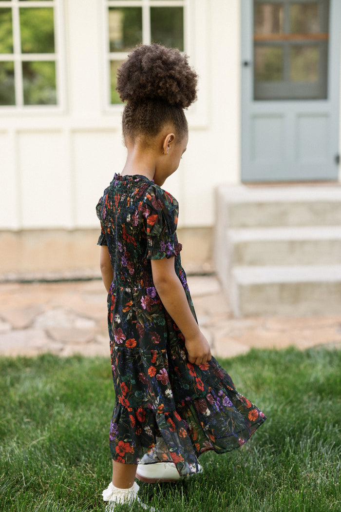 A young girl wearing the Ivy City Co Mini Cordelia Dress in Evergreen Vivid Meadow—with puff sleeves and a smocked bodice—stands on the grass, looking down. Behind her is a house with white siding, large windows, and a light blue door with stone steps.