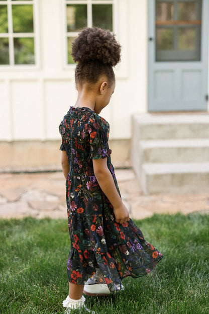 A young girl wearing the Ivy City Co Mini Cordelia Dress in Evergreen Vivid Meadow—with puff sleeves and a smocked bodice—stands on the grass, looking down. Behind her is a house with white siding, large windows, and a light blue door with stone steps.