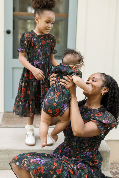 A woman and two young girls, all dressed in matching dark floral dresses with smocked bodices and playful puff sleeves, sit together on outdoor steps. The older girl stands nearby in her Ivy City Co Mini Cordelia Dress in Evergreen Vivid Meadow, watching as the woman lifts the baby with a smile.