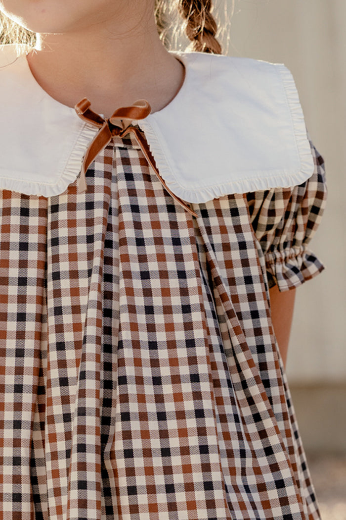A child wearing the Mini Emery Dress in Chestnut Gingham by Ivy City Co—a brown and white checked cotton dress featuring a large white collar and a small brown bow at the neckline. The puff sleeve design is complemented by the child's braided hair, with only her upper body visible.