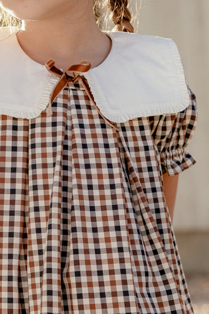 A child wearing the Mini Emery Dress in Chestnut Gingham by Ivy City Co—a brown and white checked cotton dress featuring a large white collar and a small brown bow at the neckline. The puff sleeve design is complemented by the child's braided hair, with only her upper body visible.