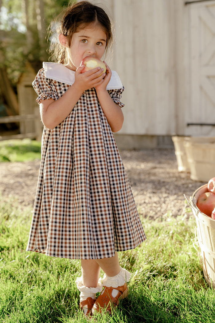 A young girl in the Mini Emery Dress in Chestnut Gingham by Ivy City Co—a charming girls’ cotton dress with puff sleeves—stands on the grass, holding and eating an apple. Nearby rests a basket of apples, with sunlight streaming over a rustic building in the background.