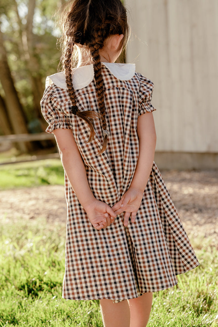 A young girl with braided hair stands outside, facing away, wearing the Ivy City Co Mini Emery Dress in Chestnut Gingham with a white collar and holding her hands behind her back. Sunlight shines on the grass and a wooden structure in the background.