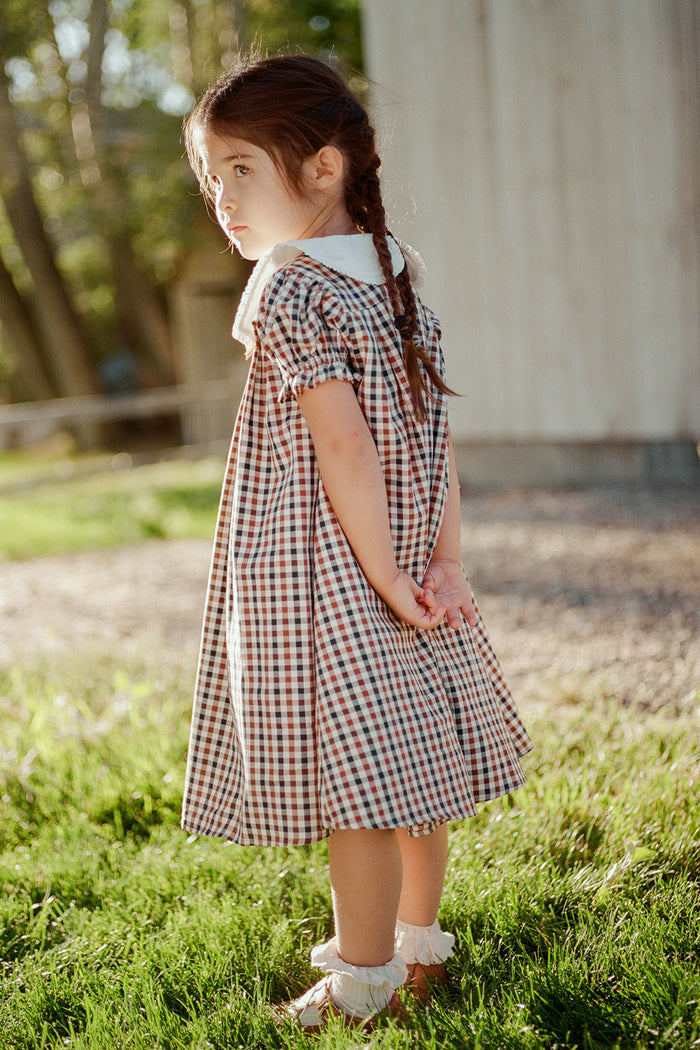 A young girl with braided hair stands on green grass in the sunlight, wearing the Mini Emery Dress in Chestnut Gingham by Ivy City Co, featuring a white collar and bows on her shoes. She looks over her shoulder with her hands behind her back, with a wooden building in the background.