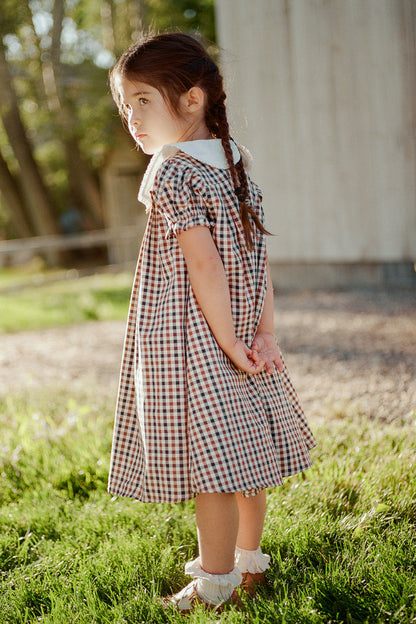 A young girl with braided hair stands on green grass in the sunlight, wearing the Mini Emery Dress in Chestnut Gingham by Ivy City Co, featuring a white collar and bows on her shoes. She looks over her shoulder with her hands behind her back, with a wooden building in the background.