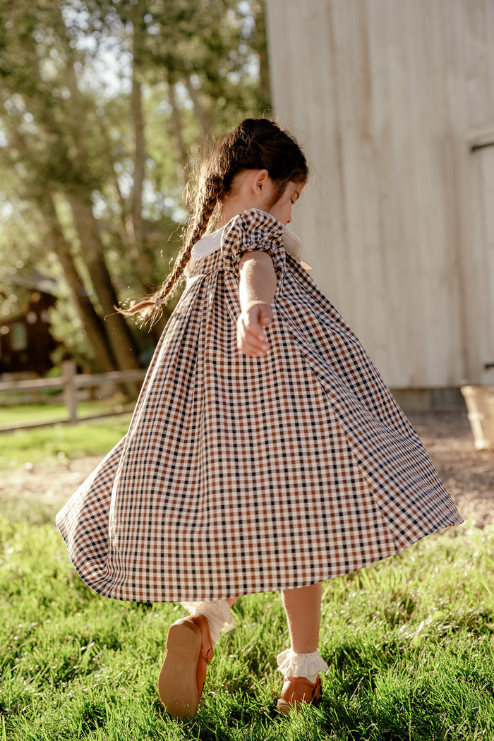 A young girl in the Ivy City Co Mini Emery Dress in Chestnut Gingham twirls on green grass, sunlight streaming through the trees behind her. Her hair is styled in braids as she stands near a wooden building.
