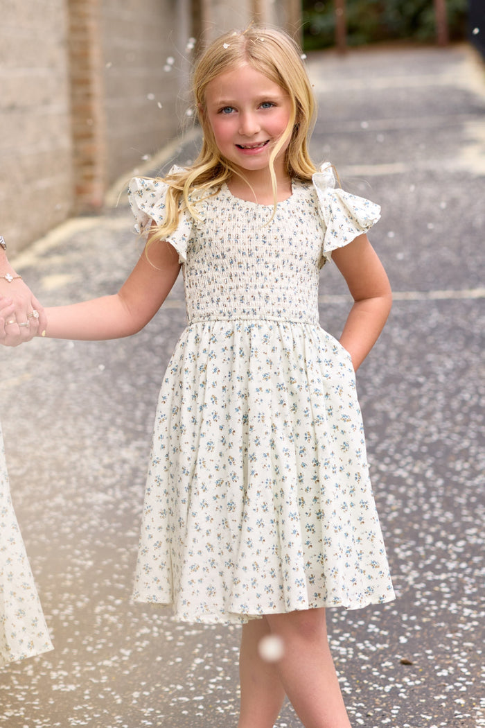 A young blonde girl stands outdoors, smiling and holding someone's hand as petals float around her, wearing the Ivy City Co Mini Hattie Dress in Madeline Blue.
