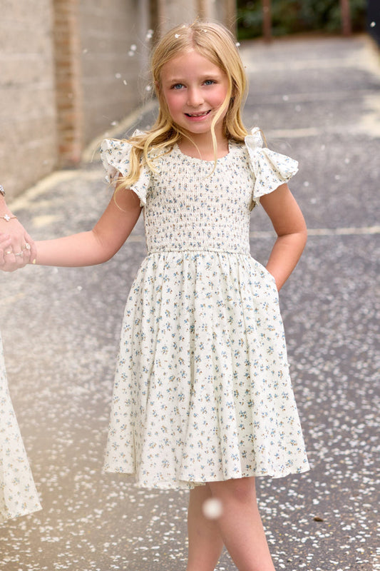 A young blonde girl stands outdoors, smiling and holding someone's hand as petals float around her, wearing the Ivy City Co Mini Hattie Dress in Madeline Blue.