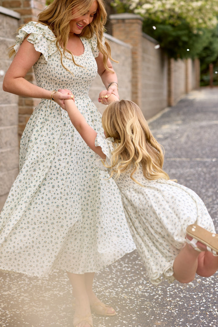 A woman and young girl smile joyfully as the woman swings the girl by her arms on a tree-lined path, both wearing Ivy City Co's matching Mini Hattie Dress in Madeline Blue.