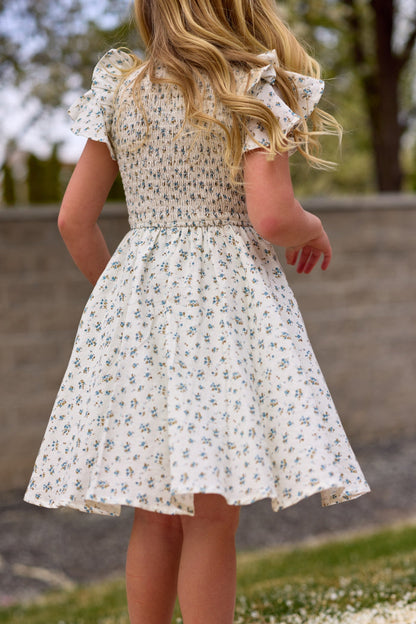 A young girl with long blonde hair stands outdoors with her back to the camera, wearing the Ivy City Co Mini Hattie Dress in Madeline Blue. Trees and a stone wall appear in the background.