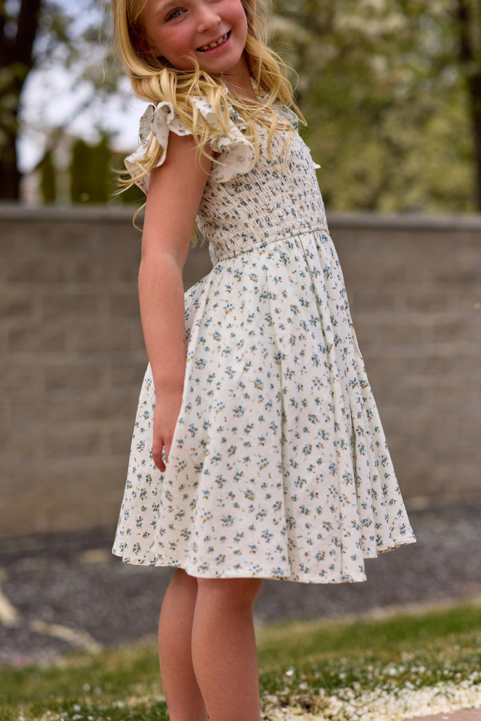 A young girl with long blonde hair smiles outdoors in the Ivy City Co Mini Hattie Dress in Madeline Blue, featuring a light blue floral pattern. She stands slightly turned, with a blurred stone wall and greenery behind her.