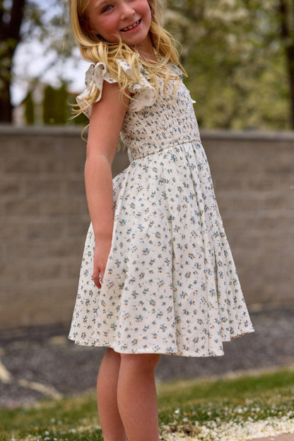 A young girl with long blonde hair smiles outdoors in the Ivy City Co Mini Hattie Dress in Madeline Blue, featuring a light blue floral pattern. She stands slightly turned, with a blurred stone wall and greenery behind her.