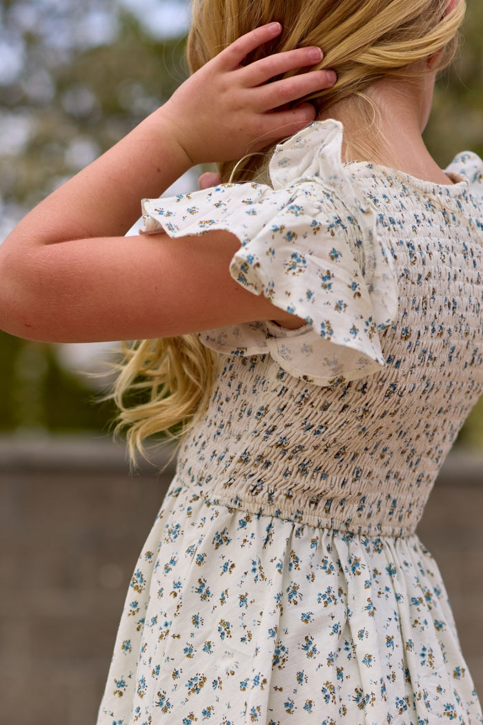 A young girl with long blonde hair wears the Mini Hattie Dress in Madeline Blue by Ivy City Co, standing outdoors with her back to the camera and touching her hair. Trees and a blurred stone wall are visible in the background.