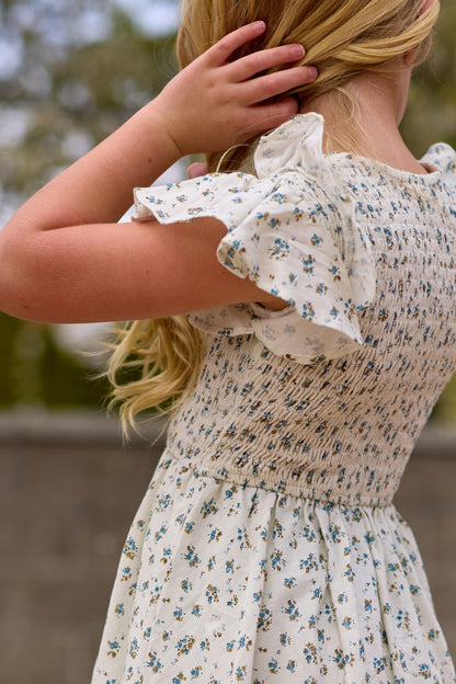 A young girl with long blonde hair wears the Mini Hattie Dress in Madeline Blue by Ivy City Co, standing outdoors with her back to the camera and touching her hair. Trees and a blurred stone wall are visible in the background.