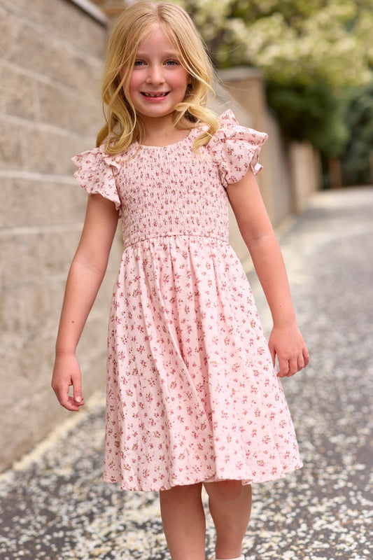 A young girl in Ivy City Co's Mini Hattie Dress in Madeline Pink—a light pink, floral girls' cotton dress—stands smiling on a paved path, with a stone wall and greenery in the background.