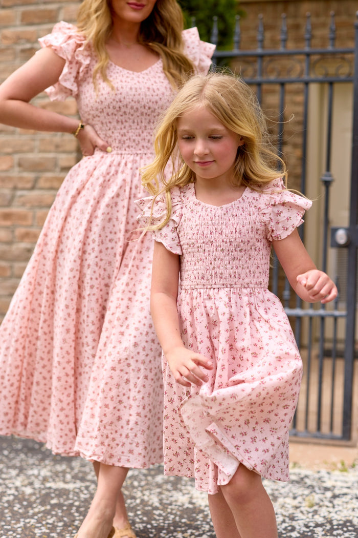 A woman and a young girl, both in matching Ivy City Co Madeline Pink floral dresses, stand outdoors by a brick wall and iron gate. The girl, wearing the Mini Hattie Dress in Madeline Pink, smiles and walks ahead.