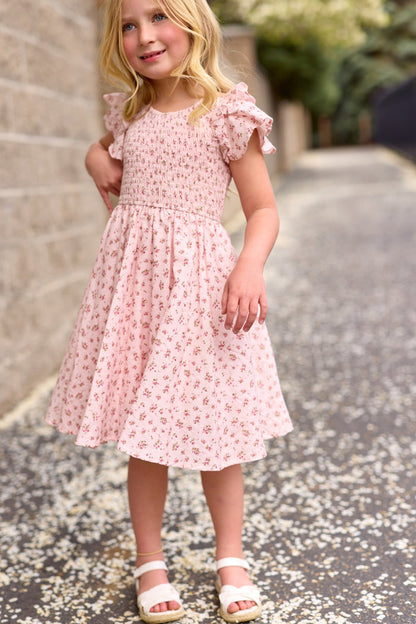 A young blonde girl wears the Ivy City Co Mini Hattie Dress in Madeline Pink, a girls’ cotton dress, while standing on a petal-strewn path and smiling with one hand on her hip.