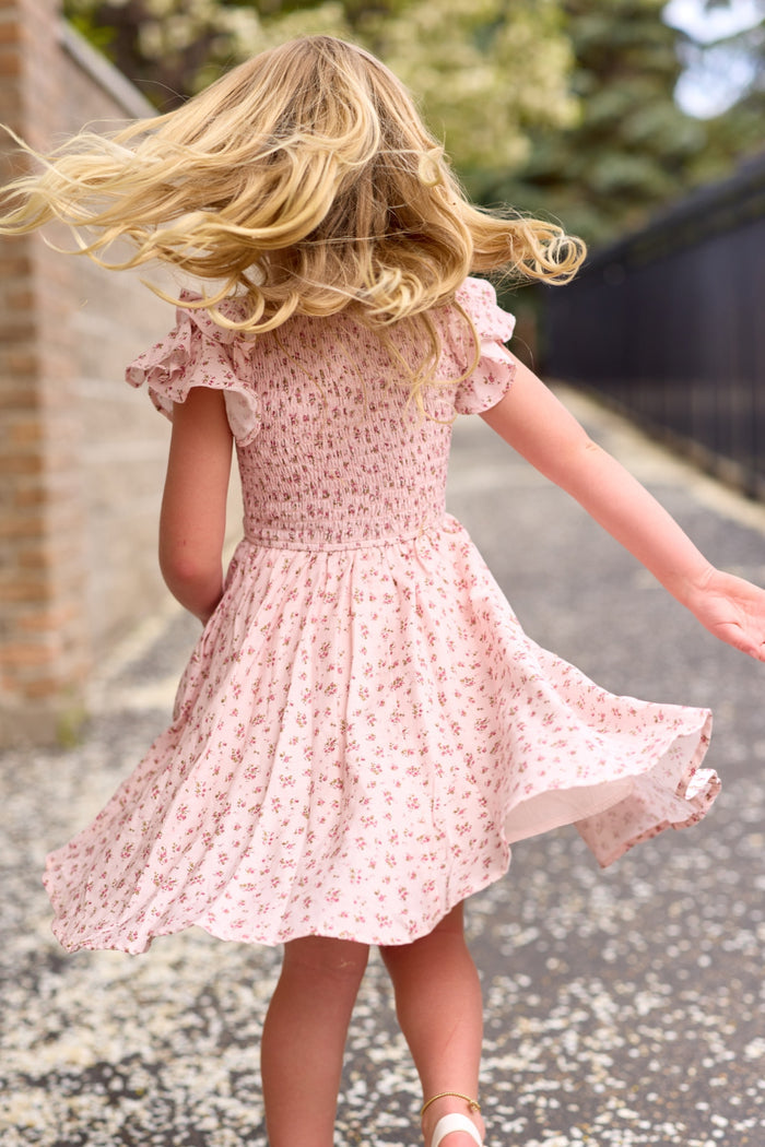 A young blonde girl twirls outdoors in the Ivy City Co Mini Hattie Dress in Madeline Pink. Viewed from behind, her hair and the floral dress flow as she spins along a paved path beside a brick wall.