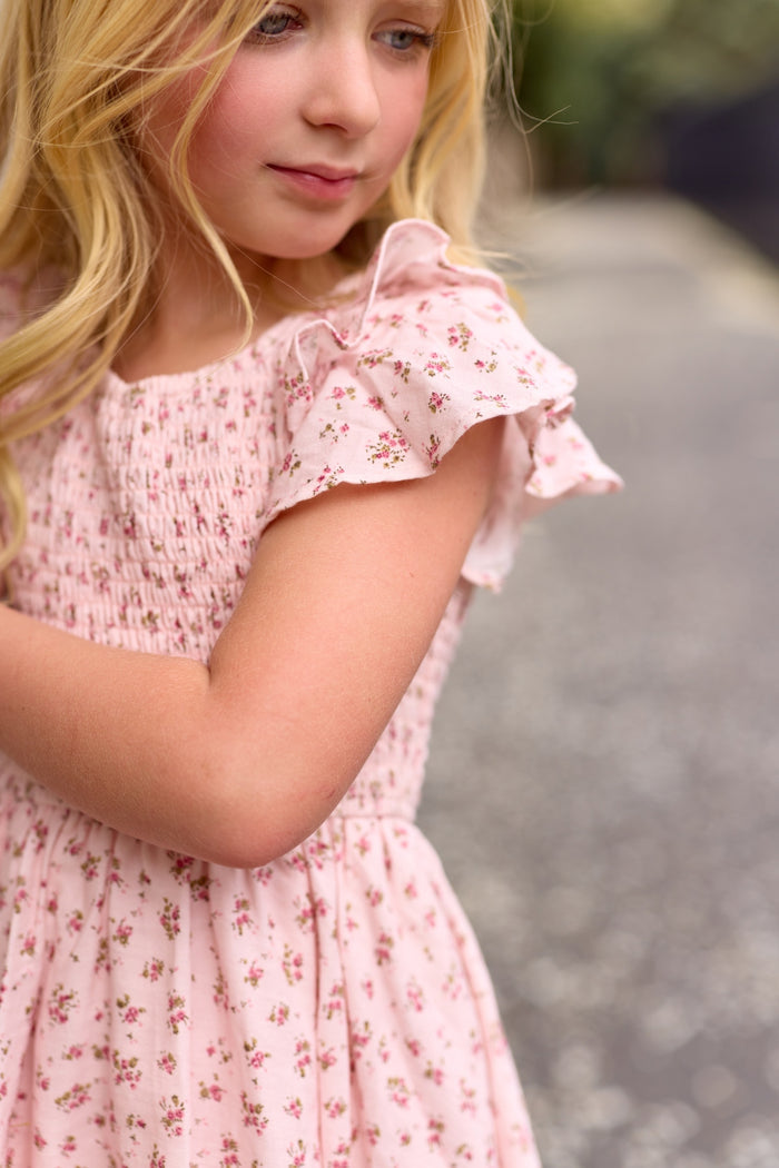 A young girl with long blonde hair wears the Ivy City Co Mini Hattie Dress in Madeline Pink, featuring ruffled sleeves, as she stands outdoors on a blurred stone path, looking down with a gentle expression.