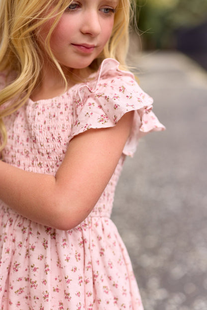 A young girl with long blonde hair wears the Ivy City Co Mini Hattie Dress in Madeline Pink, featuring ruffled sleeves, as she stands outdoors on a blurred stone path, looking down with a gentle expression.