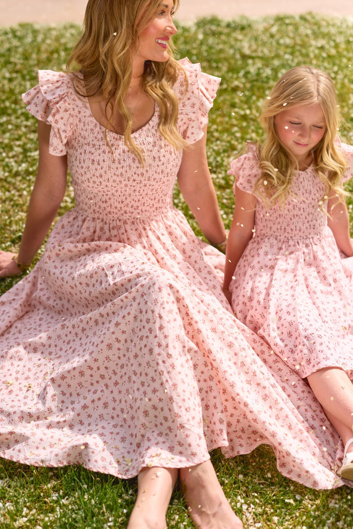 A woman and a young girl sit on grass, smiling in matching Ivy City Co Madeline Pink dresses surrounded by white flowers—the girl's outfit is the charming Mini Hattie Dress in Madeline Pink.