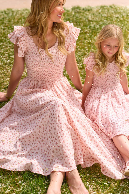 A woman and a young girl sit on grass, smiling in matching Ivy City Co Madeline Pink dresses surrounded by white flowers—the girl's outfit is the charming Mini Hattie Dress in Madeline Pink.