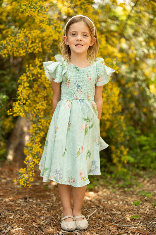 A young girl smiles outdoors on a dirt path, wearing the Ivy City Co Mini Hattie Dress in Roselyn Blue and a matching headband, standing in front of yellow flowering bushes and greenery.