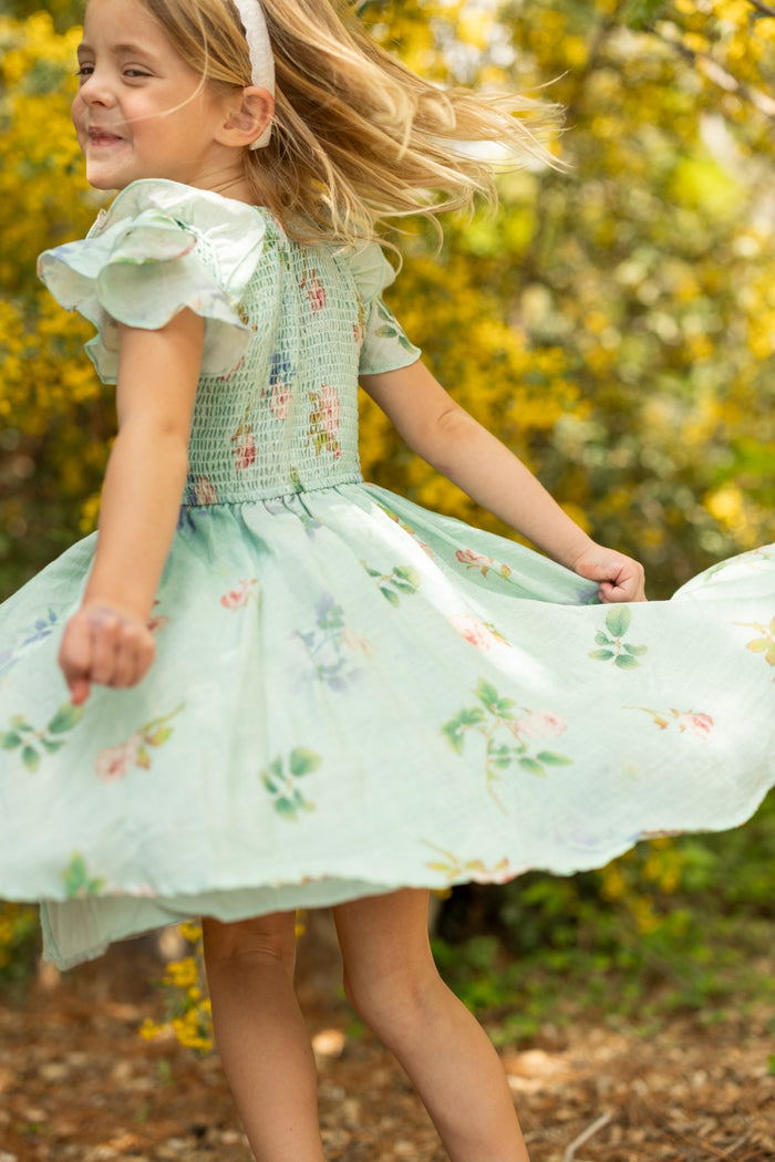 A young girl with blonde hair wearing the Ivy City Co Mini Hattie Dress in Roselyn Blue and a white headband twirls joyfully outdoors, surrounded by yellow flowers and greenery in bright sunlight.