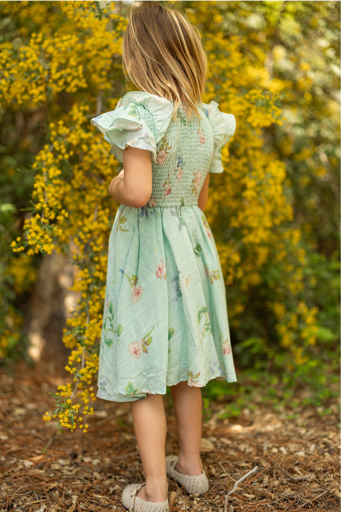 A young girl in Ivy City Co’s Mini Hattie Dress in Roselyn Blue stands outdoors with her back to the camera, facing bright yellow flowering bushes. She has light brown hair and stands on a path covered with dry leaves.