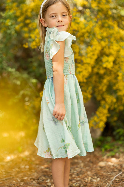 A young girl with light brown hair, wearing the Ivy City Co Mini Hattie Dress in Roselyn Blue and a white headband, stands outdoors near yellow flowering bushes, looking at the camera with a slight smile.