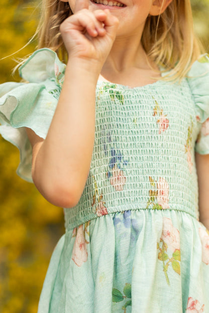 A young girl smiles in a partially visible pose, wearing the Ivy City Co Mini Hattie Dress in Roselyn Blue with ruffled sleeves. She holds up her hand against a warmly blurred yellow background.