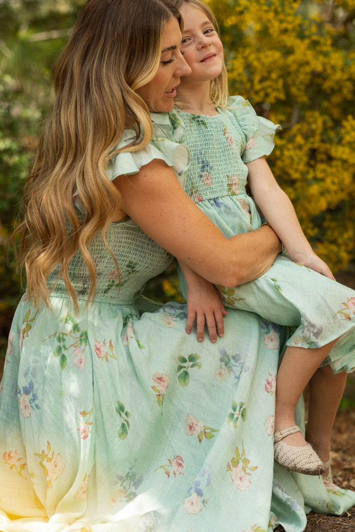 A woman with long wavy hair holds a young girl on her lap. Both wear matching Ivy City Co Mini Hattie Dresses in Roselyn Blue. Outdoors among yellow flowers, they share a warm and affectionate moment.